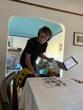 Seth Young standing and leaning forward, with headphones on, reaching into a bag with one hand and holding a laptop in the other